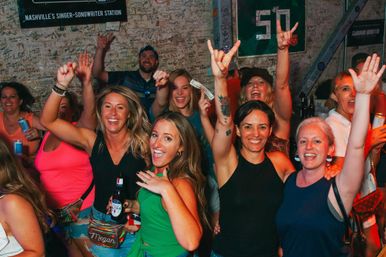 Energetic group of people cheering and holding drinks at a crowded Nashville live-music bar, smiling faces and raised hands against a graffiti-covered wall.
