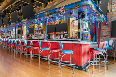 Retro-style urban bar interior with a long red counter, a row of red-and-blue vinyl stools, colorful string lights, multiple flat-screen TVs, hardwood floor and exposed brick wall.