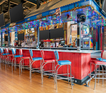 Retro-style urban bar interior with a long red counter, a row of red-and-blue vinyl stools, colorful string lights, multiple flat-screen TVs, hardwood floor and exposed brick wall.