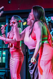 Three women singing into microphones on an indoor nightclub stage with vibrant pink and green lighting and bright dresses, lively live-music performance.