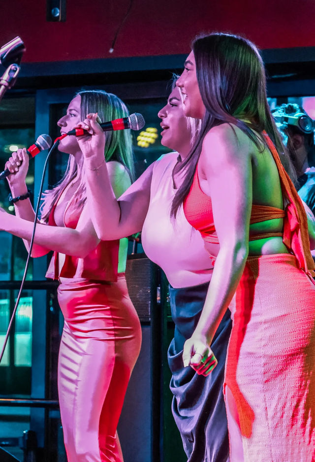 Three women singing into microphones on an indoor nightclub stage with vibrant pink and green lighting and bright dresses, lively live-music performance.