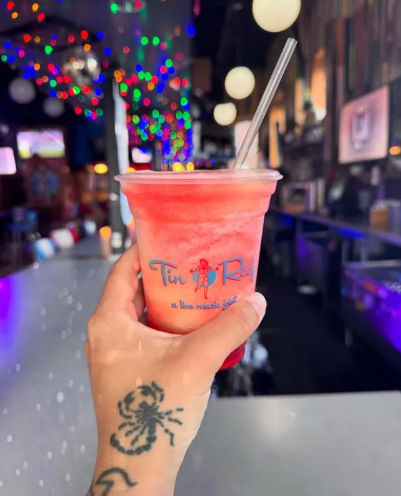 Hand with scorpion tattoo holding a bright pink frozen drink in a plastic cup with a straw at a lively bar counter, colorful bokeh string lights in the background