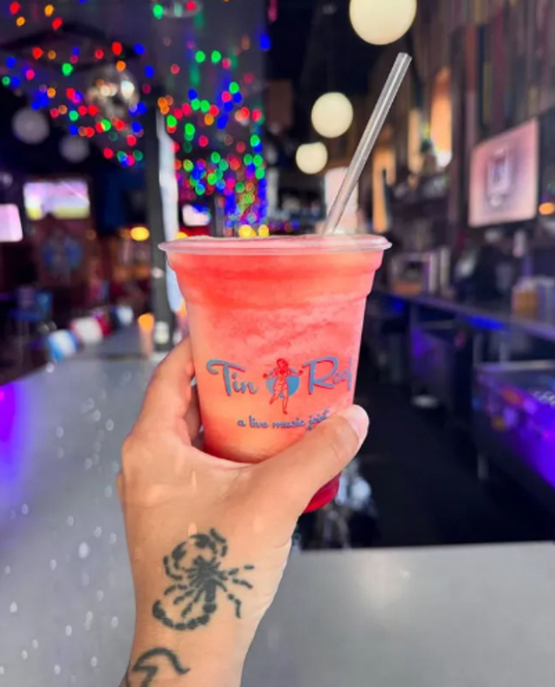 Hand with scorpion tattoo holding a bright pink frozen drink in a plastic cup with a straw at a lively bar counter, colorful bokeh string lights in the background