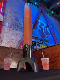 Neon-lit bar interior with a tall illuminated drink tower of pink slushy on a table between two plastic cups with straws, graffiti-covered walls and colorful string lights.