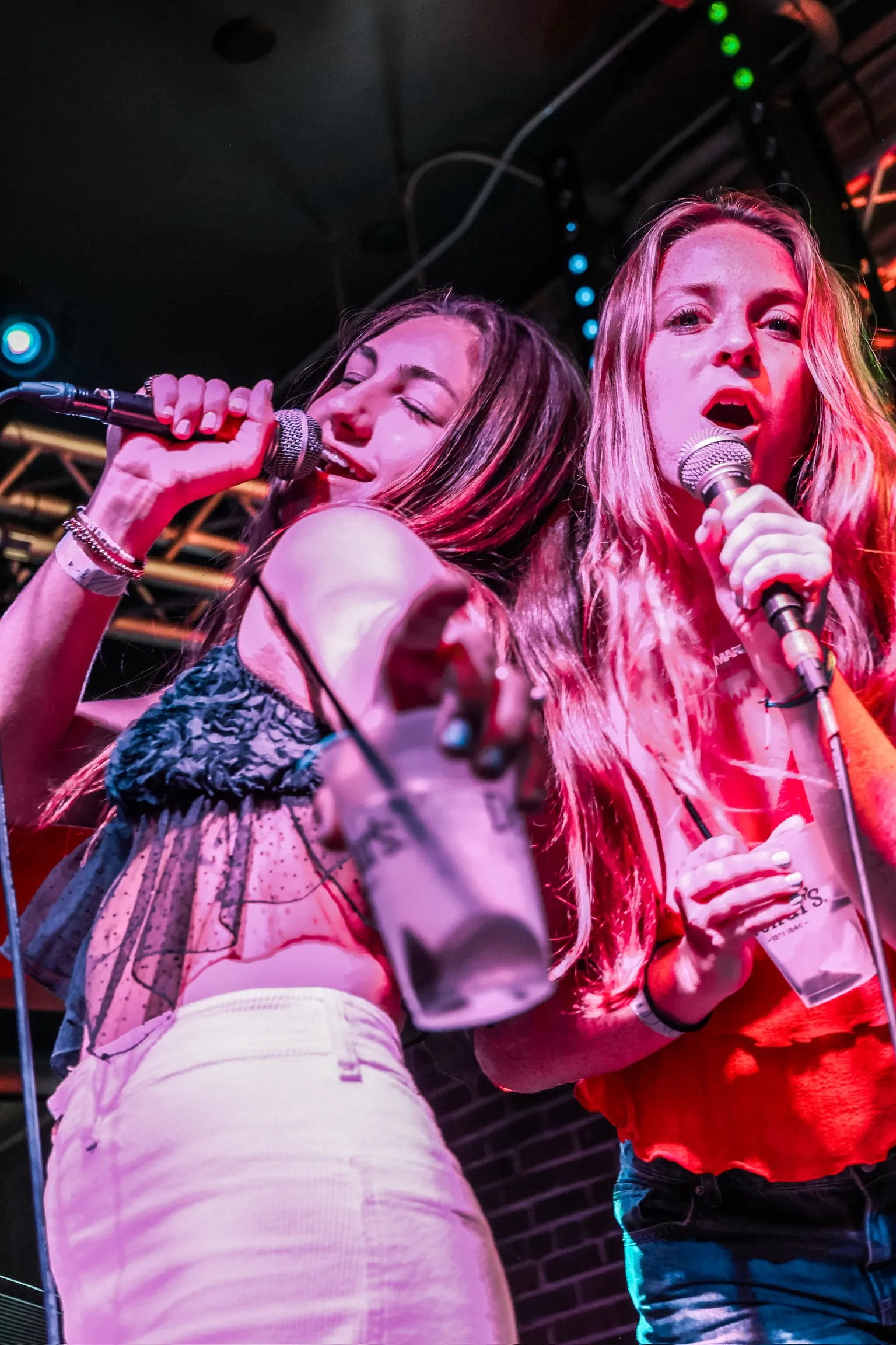 Two performers singing a duet on a neon-lit bar stage, each holding a microphone and a plastic cup toward the crowd during a lively live-music night