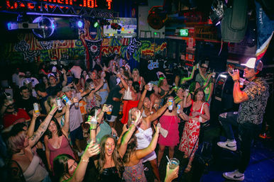 Lively indoor bar scene with colorful murals and neon lights, a packed crowd of partygoers raising cups and cans toward a male singer on a small stage under a disco ball.