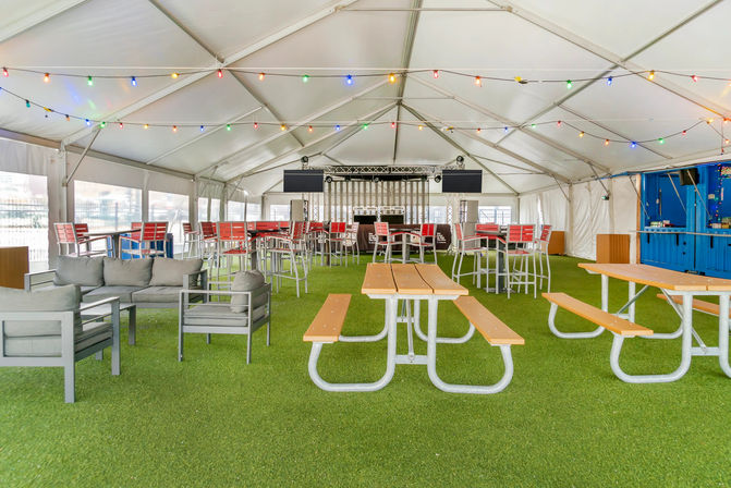 Large outdoor event tent with artificial turf, picnic tables and benches, gray lounge seating, red high-top chairs around a central bar, stage and speakers at the far end, and colorful string lights overhead.