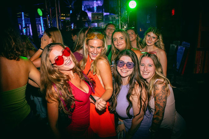 Friends posing at a lively nightclub dance floor, wearing colorful dresses and sunglasses, smiling under green and purple stage lights with a DJ booth in the background.