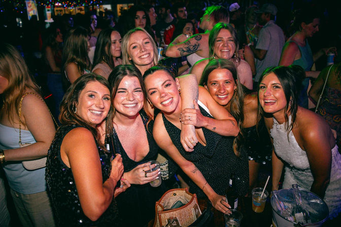 Energetic group of friends laughing and posing with cocktails at a crowded nightclub bar under colorful string lights, dancing and hugging for a night out