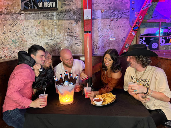 Five friends laughing in a neon-lit dive bar booth, sharing an illuminated bucket of beer bottles and a tall beer tower with plastic cups, chips and salsa against graffiti-covered walls.