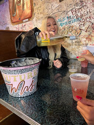 Woman pouring white wine into plastic cups at a lively bachelorette party in a graffiti-covered bar booth, ice bucket reading 'Future Mrs.'