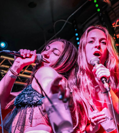 Two women singing into microphones during a live club performance, bathed in pink and blue stage lighting.