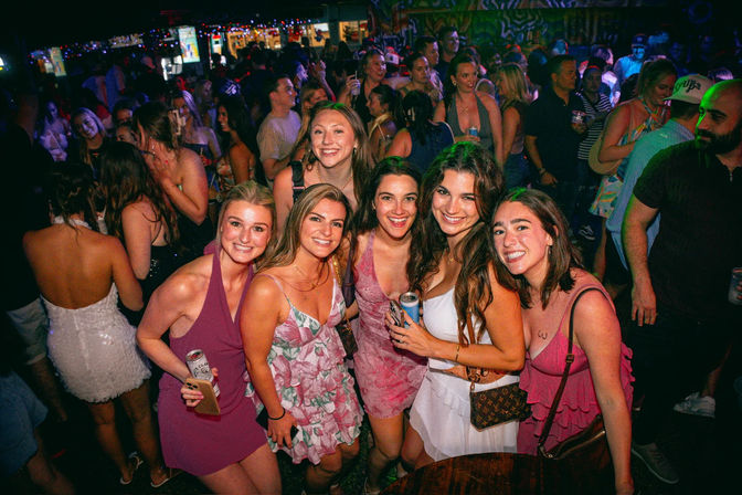 Group of young adults smiling and posing with drinks on a crowded, colorful nightclub dance floor — lively bar nightlife scene with friends partying