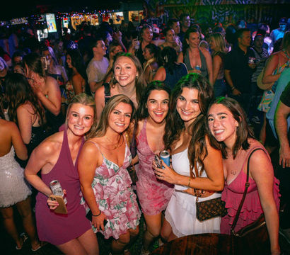 Six smiling women in summer dresses posing with drinks at a packed, colorful nightclub or bar, festive lights and a lively crowd dancing in the background.