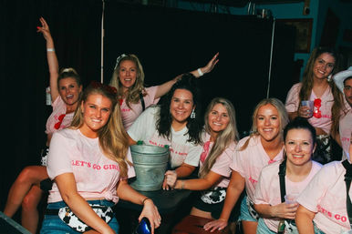Cheerful group of women in matching pink "Let’s Go Girls" shirts posing around a table with a metal drink bucket at a lively bar, smiling and holding cups.