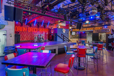 Colorful live music venue interior with neon "Drink Dance Repeat" sign, stage with drum kit and disco ball, retro red-and-blue diner-style chairs and bar tables.
