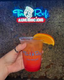 Hand holding a red-to-orange layered cocktail in a logoed plastic cup with ice and an orange slice garnish, neon live-music sign glowing blurred against a textured wall in the background.