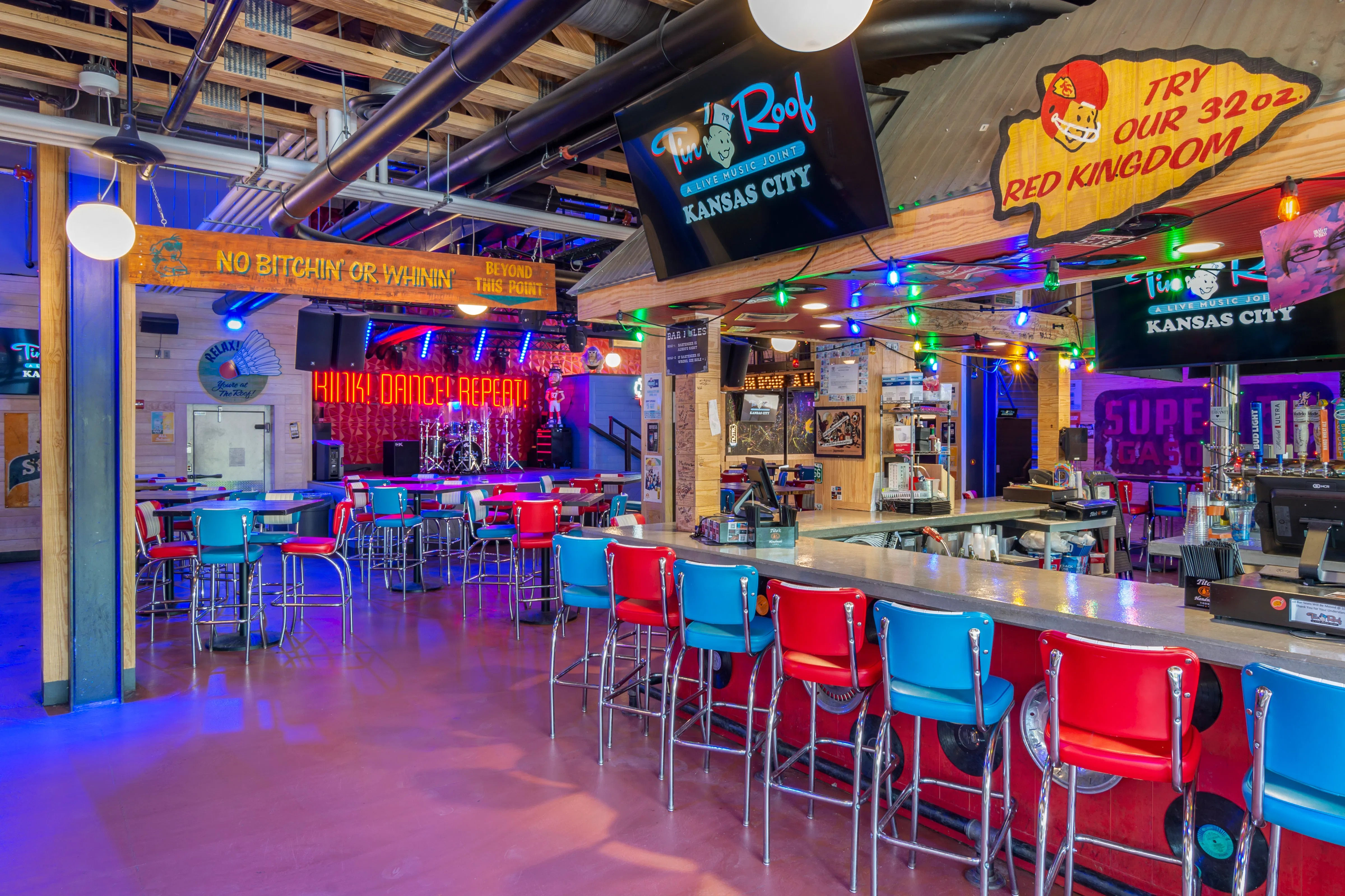 Vibrant Kansas City live-music bar interior with retro red and blue diner stools at a long counter, small stage with drum kit and neon dance-repeat sign, exposed industrial ceiling and colorful string lights