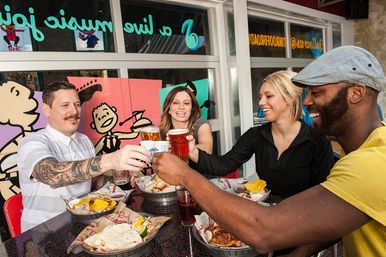 Four friends toasting pints on a colorful casual restaurant patio, sharing burgers, nachos and fries in a cheerful outdoor dining scene.