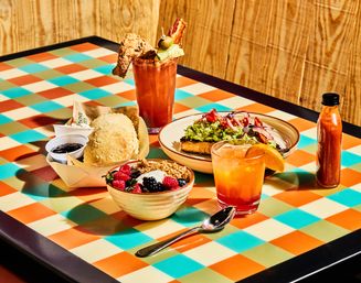 Colorful checkered-table brunch: avocado toast with pickled onions and cherry tomatoes, a bowl of fresh berries and granola, a flaky biscuit with jam, a bacon-topped Bloody Mary, an orange cocktail with orange slice, and a bottle of hot sauce against a wood-paneled backdrop.