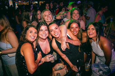 Smiling group of friends posing with drinks at a crowded bar/nightclub with colorful lights — fun city nightlife scene.