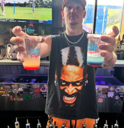 Bartender in a sleeveless graphic shirt holding two colorful layered cocktails at a lively bar counter with sports on TV and sticker-covered taps