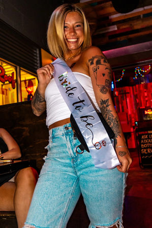 Smiling woman wearing a 'bride to be' sash over a white tube top, ripped light-blue jeans and visible tattoos, posing in a colorful neon-lit bar for a bachelorette party.