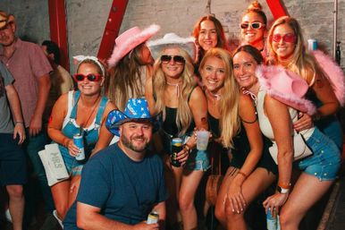 Smiling group of friends at an indoor bar party, women in pink cowboy hats, feather boas and sunglasses with denim shorts, holding drinks against a brick wall