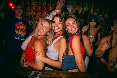 Three smiling women hugging and posing at a crowded urban nightclub bar with colorful graffiti walls, bright party lights and people dancing in the background — lively nightlife scene.