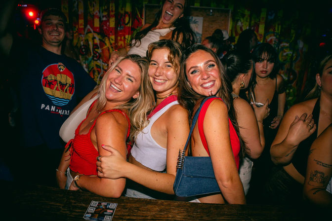 Three smiling women hugging and posing at a crowded urban nightclub bar with colorful graffiti walls, bright party lights and people dancing in the background — lively nightlife scene.