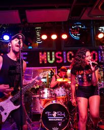 Energetic live band on a neon-lit indoor stage — male guitarist singing into a mic, female lead vocalist belting lines, and drummer behind a sparkling kit under colorful marquee lights, lively nightlife performance.