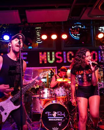 Energetic live band on a neon-lit indoor stage — male guitarist singing into a mic, female lead vocalist belting lines, and drummer behind a sparkling kit under colorful marquee lights, lively nightlife performance.