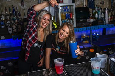 Two bartenders laughing as one pours a stream of liquor into a blue cocktail shaker over the bar counter with neon-blue backlighting and plastic drink cups.