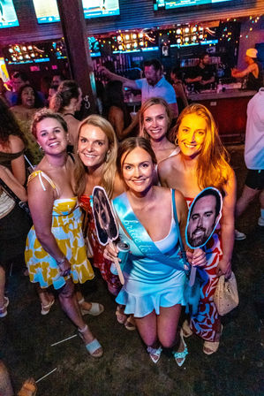 Smiling group of women at a neon-lit crowded bar celebrating a bachelorette party; bride-to-be wearing a light-blue sash holds face-cutout props while friends in colorful summer dresses pose near the busy bar and TV screens