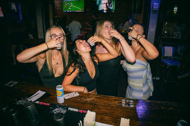 Four friends at a wooden bar taking shots together, laughing and leaning on the counter in a dim, neon-lit nightlife scene with TVs on the wall.
