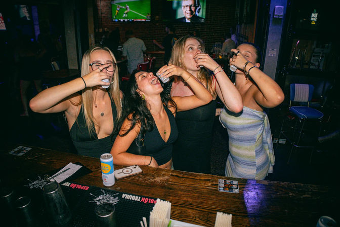 Four friends at a wooden bar taking shots together, laughing and leaning on the counter in a dim, neon-lit nightlife scene with TVs on the wall.