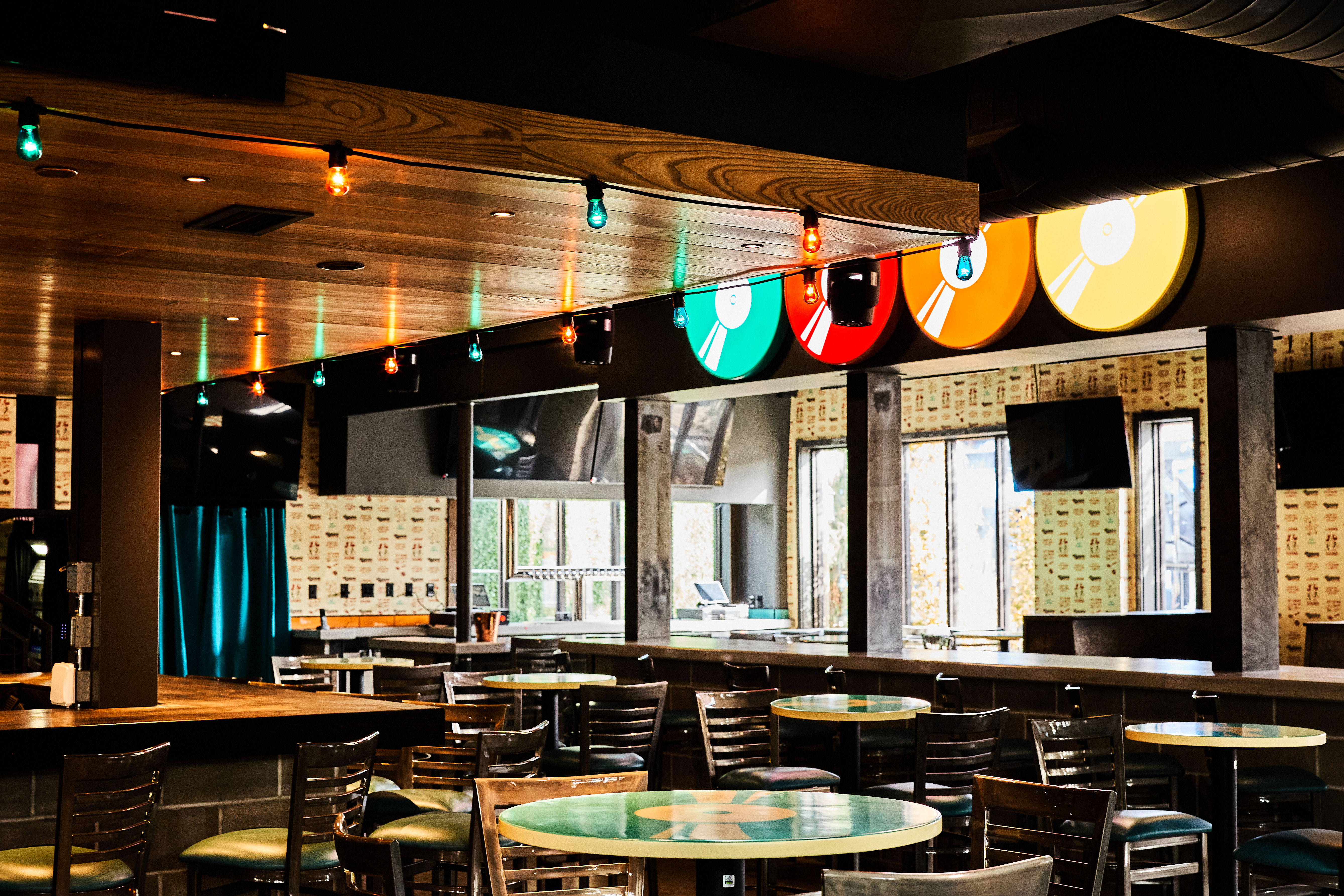 Empty modern bar interior with wooden ceiling, colorful vinyl-record themed signs and hanging string lights, round tables and high stools, large windows and wall-mounted TVs — casual taproom/lounge vibe.