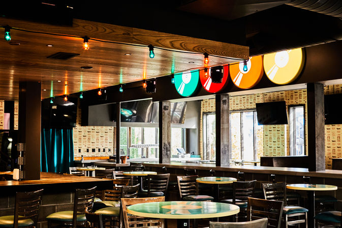 Empty modern bar interior with wooden ceiling, colorful vinyl-record themed signs and hanging string lights, round tables and high stools, large windows and wall-mounted TVs — casual taproom/lounge vibe.