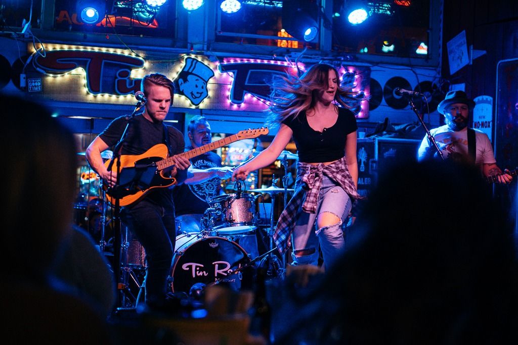 Energetic live band performing on a neon-lit bar stage with a guitarist, drummer and a dancing female singer in ripped jeans, audience silhouettes in the foreground.