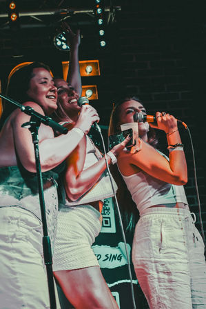 Three friends singing into microphones on a lively karaoke stage in a dimly lit bar, smiling and holding a drink and phone under colorful stage lights.