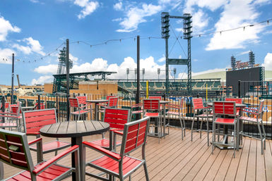Sunny rooftop deck with red patio chairs, round tables and string lights overlooking a baseball stadium and city skyline under a blue sky.