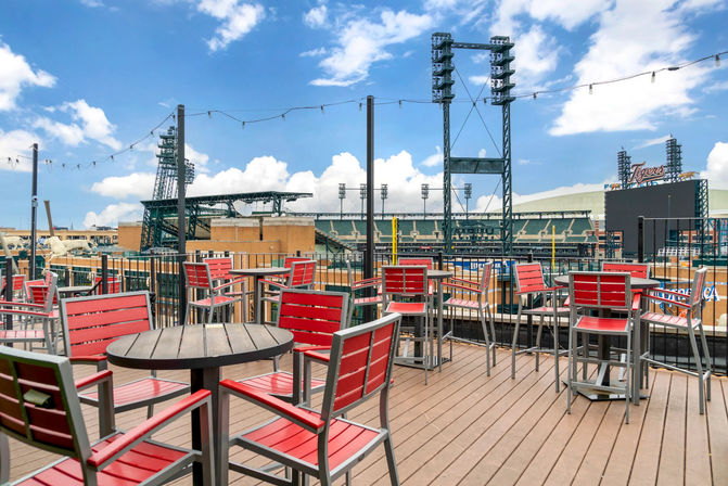 Sunny rooftop deck with red patio chairs, round tables and string lights overlooking a baseball stadium and city skyline under a blue sky.