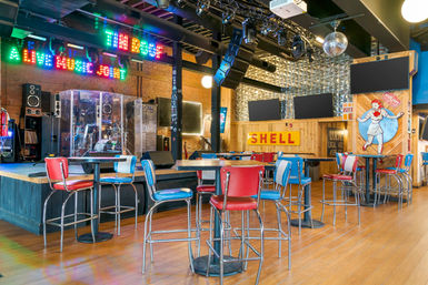 Colorful indoor live music venue with stage and drum kit behind plexiglass, neon live-music sign, retro red and blue diner-style bar stools and high-top tables, disco ball and wooden floors.