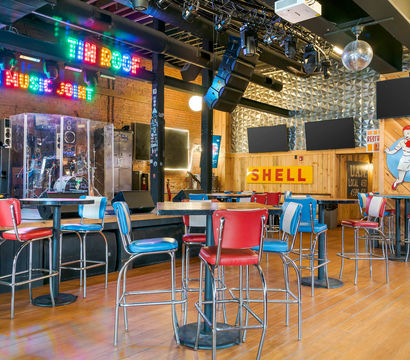 Colorful indoor live music venue with stage and drum kit behind plexiglass, neon live-music sign, retro red and blue diner-style bar stools and high-top tables, disco ball and wooden floors.