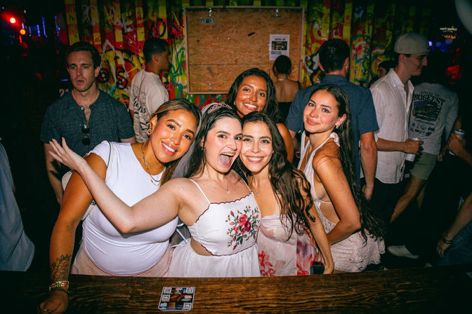 Five friends smiling and posing at a lively nightclub bar in summer dresses, colorful graffiti wall and neon-lit crowd in the background — fun nighttime party scene.