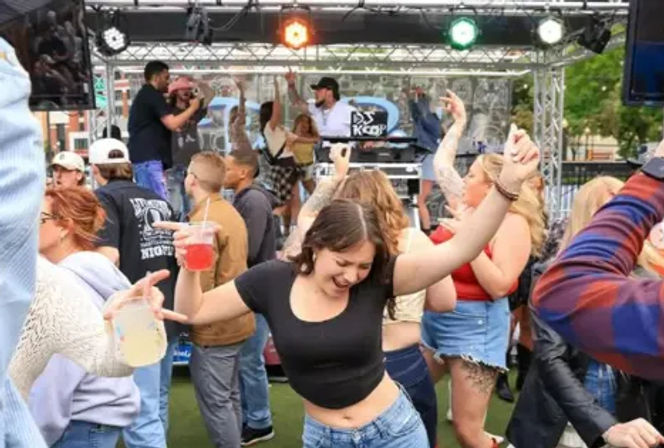 Young woman dancing with a red drink in hand amid a lively outdoor music festival crowd, DJs on an elevated stage with colored lights overhead