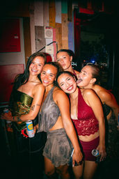Five young women smiling and posing together in a colorful bar at night, holding canned drinks and wearing party outfits — one playfully sticking out her tongue in a lively city nightlife scene.