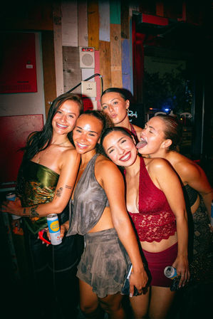 Five young women smiling and posing together in a colorful bar at night, holding canned drinks and wearing party outfits — one playfully sticking out her tongue in a lively city nightlife scene.
