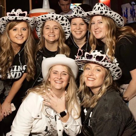 Smiling group of six women wearing cow-print and white cowboy hats at a lively bar bachelorette party, bride proudly showing her engagement ring