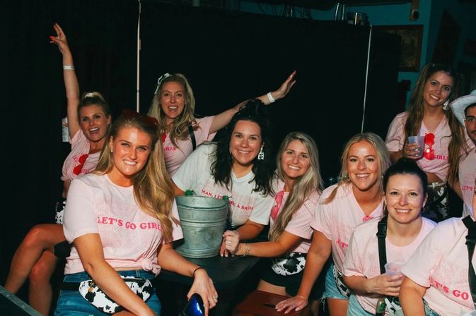 Group of smiling women in matching pink Let's Go Girls T-shirts and cow-print waist bags posing around a table with a metal bucket and drinks in a dim bar, lively girls' night out.